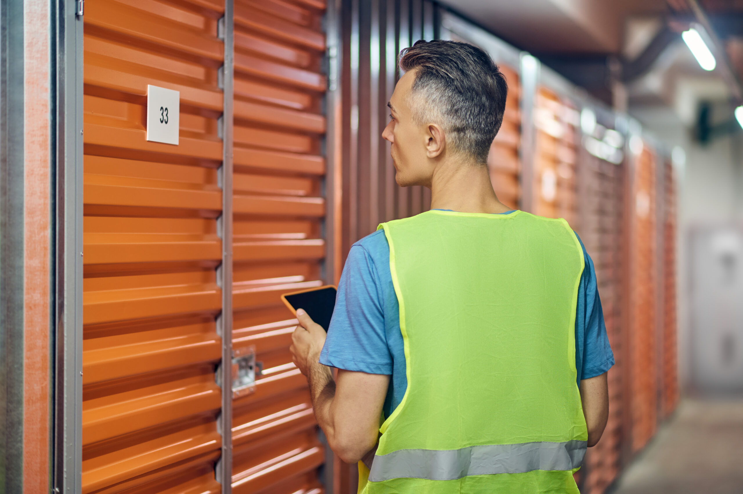 Warehouse security. Man in bright vest with tablet working in warehouse guard standing with back to camera near garage door